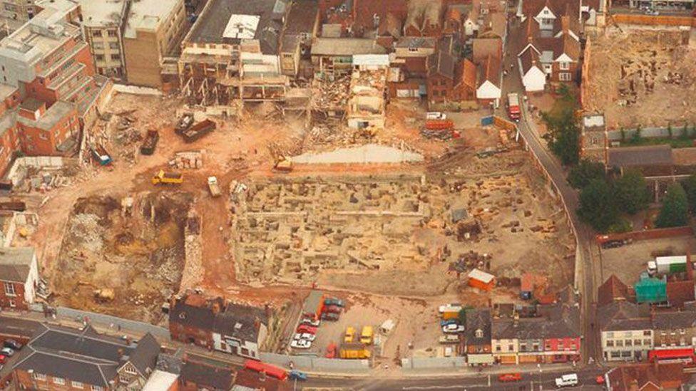 A grainy colour aerial shot of Ipswich showing an excavation in Ipswich town centre. Buildings surround a cleared site and in the middle are clear medieval foundations and archaeologists' trenches.