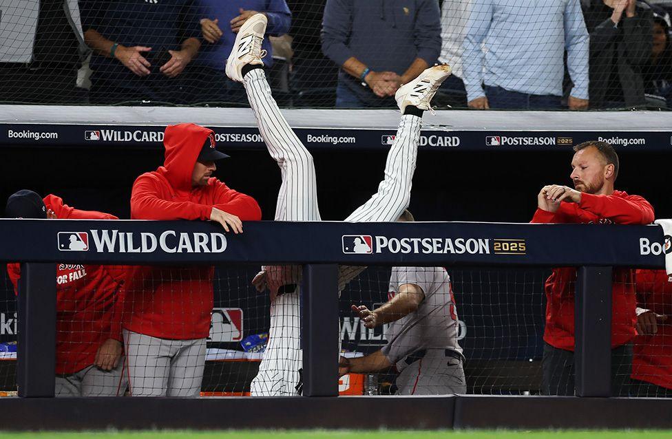 New York Yankees’ Ryan McMahon leaps over the dugout wall to catch a pop fly during the eighth inning against the Boston Red Sox at Yankee Stadium in New York City. His legs are visible above the railing, he's upside down with team mates looking on.