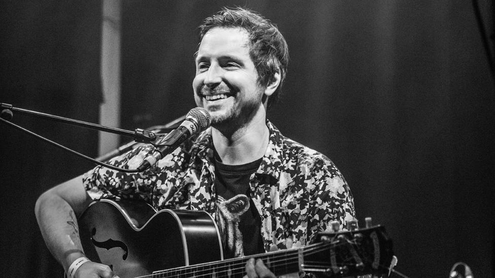 Black and white image of Ollie Thomas sat on stage with his guitar, in front of a microphone, smiling. Ollie has short hair and dark stubble. He wears a t-shirt. It looks like it might have a bear on it, though is partially covered by a short-sleeved shirt worn over it with the buttons undone.