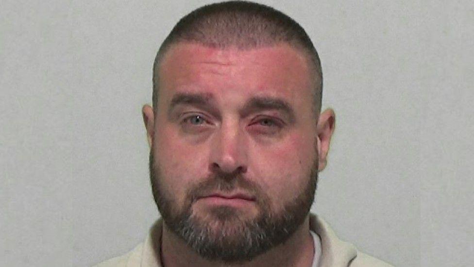 Andrew Tait is a white man with a buzz-cut hairstyle and a trimmed stubble. He squints at the camera and has an injured left eye. He has a round face and is of larger build.