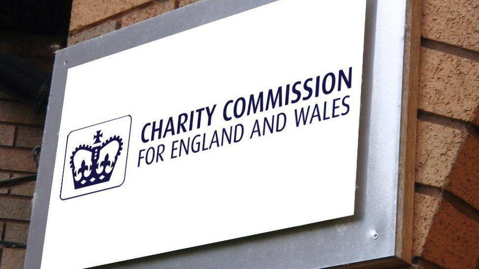 Blue and white sign showing a sign reading "Charity Commission For England and Wales" with a an image of a crown beside the wording. The sign is hung on a brick pillar.