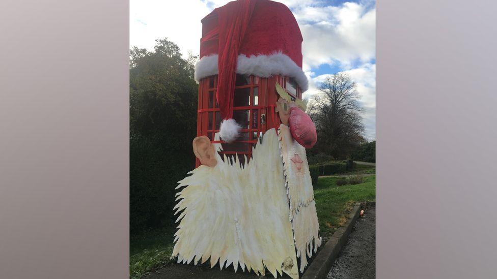 A Santa face on a telephone box. There is a red hat on the top of the box and the Santa model has large eyes, a big red nose, ears, and a beard
