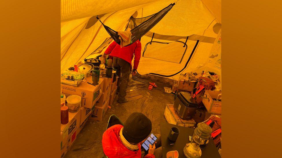 Two people are in a tent. They are wearing jackets and woolly hats and are surrounded by boxes of equipment and provisions including cooking equipment