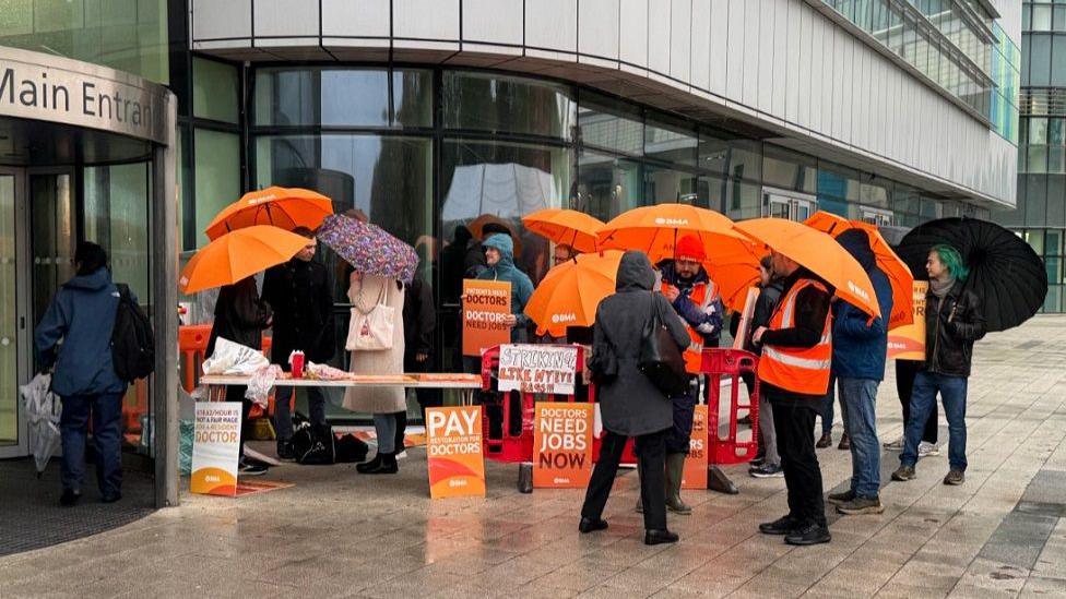 Orange umbrellas and orange signs talking about the strikes are outside the hospital, with people wearing coats with the hoods up due to the rain.