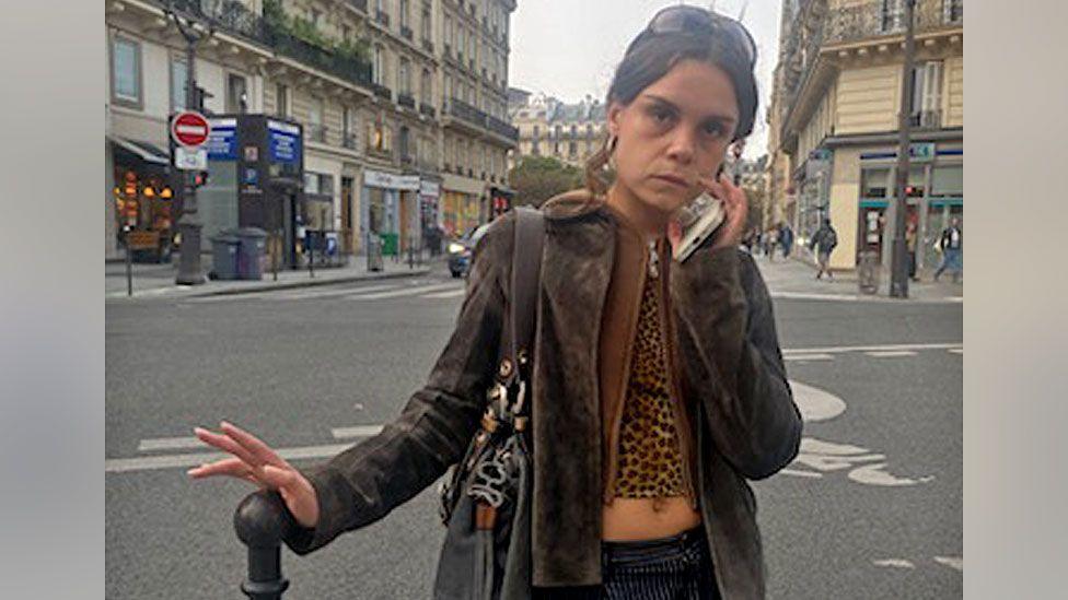 A young woman with brown hair stands in the street, resting her hand on a bollard. She is wearing a brown jacket and a leopard-print top. She is holding her phone up to her face and has a serious expression