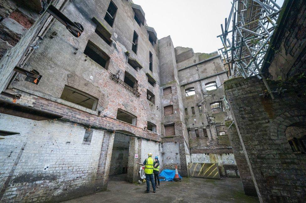 Interior courtyard of a large derelict industrial building with exposed brickwork, empty window openings and weathered concrete walls. Two construction workers wearing high-visibility clothing and hard hats stand on the ground floor inspecting the site, while extensive metal scaffolding rises along one side of the structure. The scene is lit by daylight from above, highlighting the building's deteriorated condition and multiple storeys.