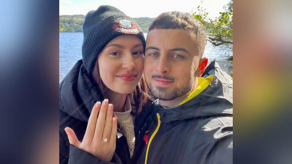 Faith is wearing a black coat and dark coloured beanie, and is holding up her hand. On her finger is an engagement ring. Next to her is her partner Sam, who has short dark brown hair and black facial hair. Behind them is a lake.