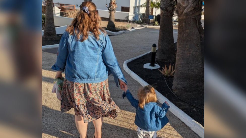 Mary and her daughter walking down the street. Maru is wearing a blue denim jacket and floral skirt. Her daughter is also wearing a blue denim jacket and a white bottoms.