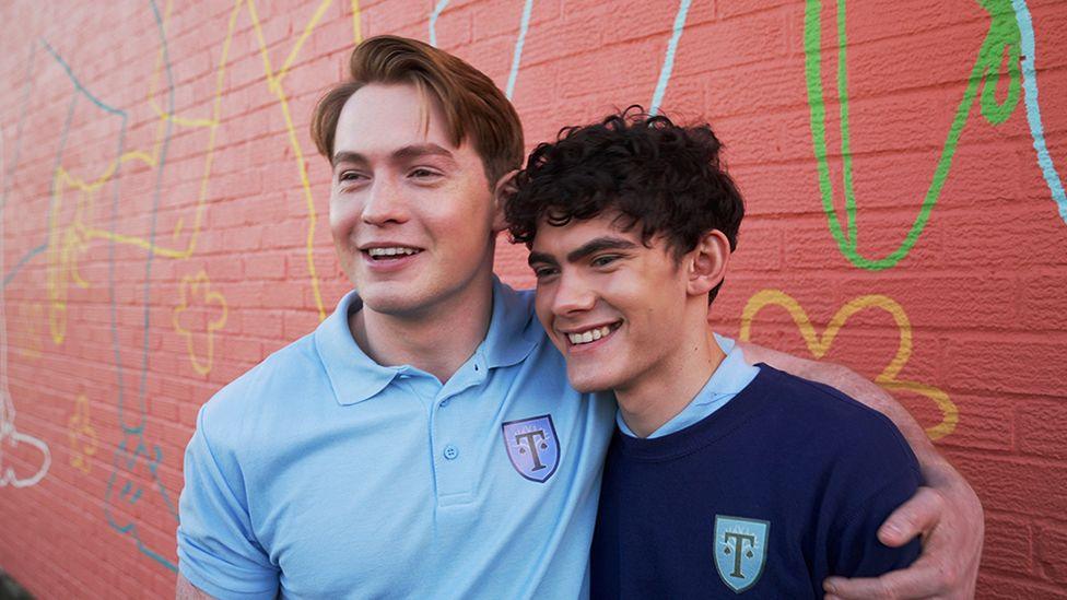 Actors Kit Connor and Joe Locke smile smile at the camera, they're both in their Truham High PE kits from the show. Kit has his arm around Joe.