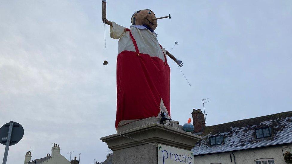 Statue with red trousers held up by red braces over a white shirt. The model head has a long protruding nose. 