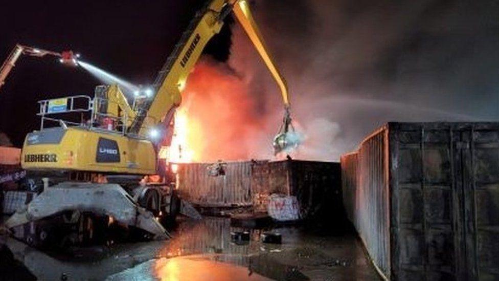 Yellow digger with arm reaching over a metal container where a large fire glows