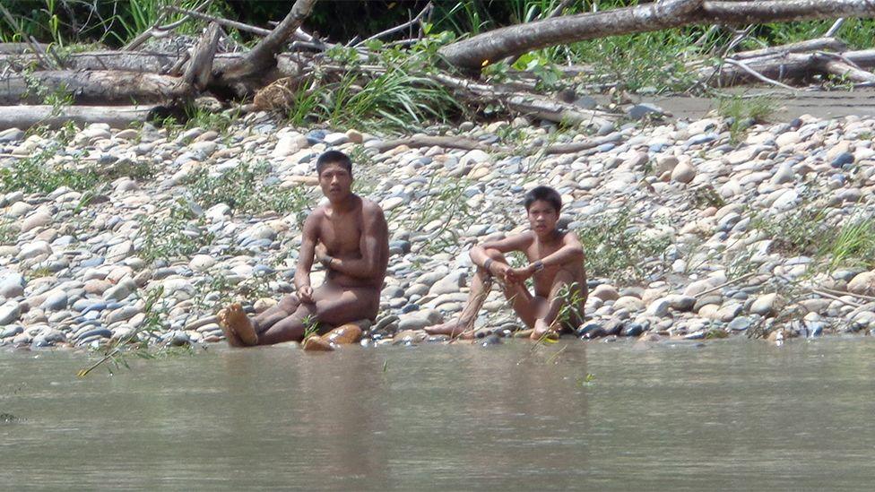 Two individuals are sitting on a rocky riverbank near the water’s edge. They appear unclothed and are positioned on smooth stones with some scattered vegetation around them. Behind them, there are fallen tree trunks and green foliage, indicating a natural, forested environment. The river in the foreground has calm, shallow water.
