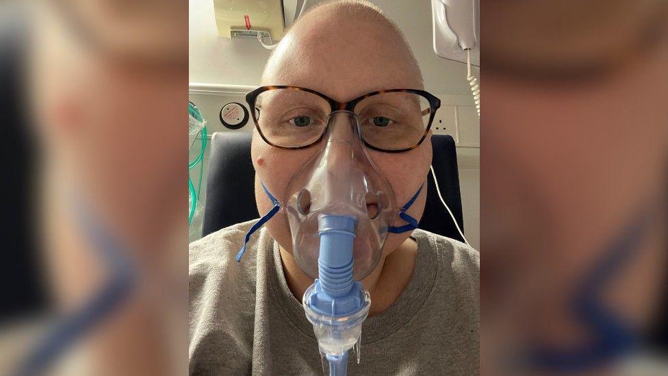 A head-and-shoulders shot of a woman with no hair wearing glasses and a oxygen mask.