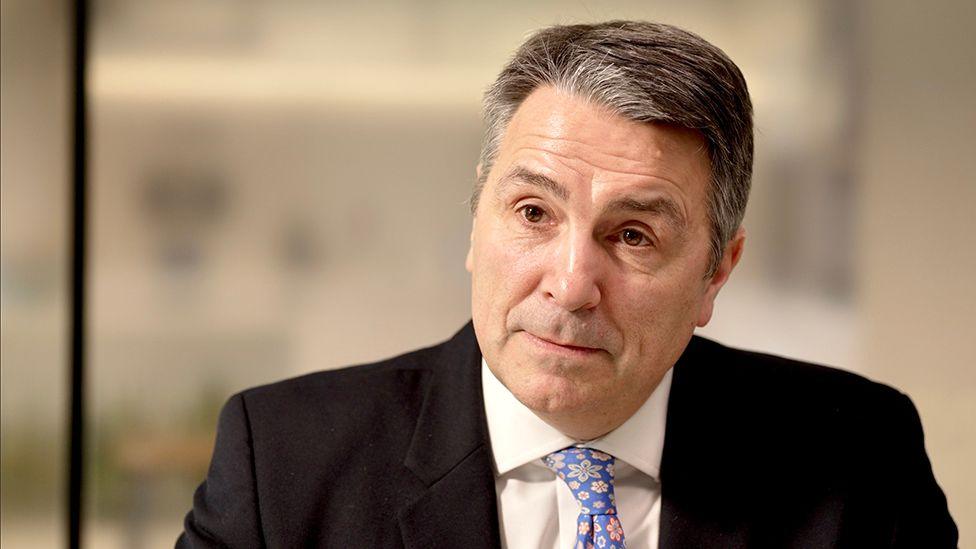 Headshot of Stephen Vullo. He has short, dark, greying hair, is clean shaven and is wearing a black suit, white shirt and a blue tie patterned with pastel-coloured flowers.