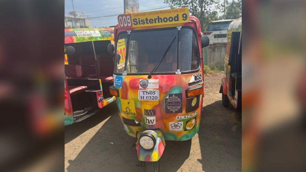 A tie-dye coloured tuk-tuk with lots of stickers from businesses stuck onto it. It has a yellow sign at the top that reads "009 Sisterhood 9", and is parked next to other tuk-tuks.