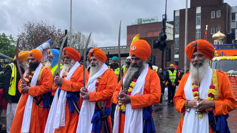 Five men in traditional orange dress holding swords vertically with crowds in the background