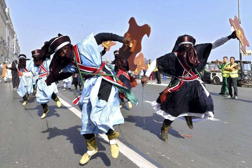 Algerian men dance in formation, waving swords ands shields.