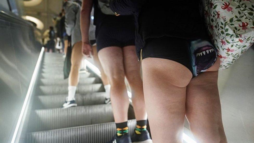 A woman wearing pants with shark jaws on them rides a station escalator with fellow event attendees