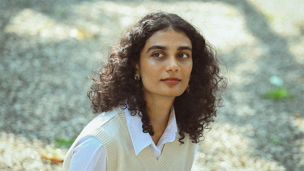 Anumita, wearing a cream coloured sweater vest over a white shirt, looking to the side while sitting outside.