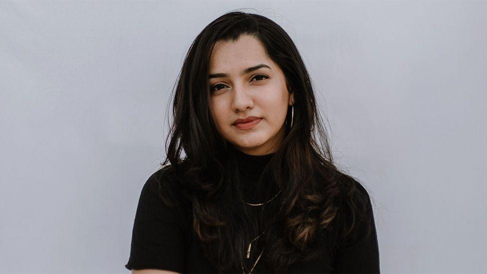 Frizzell D'Souza, wearing a black top and shiny necklace. The background is plain light grey.