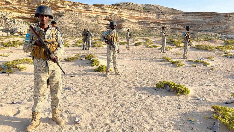 Puntland Maritime Police Force officers standing in uniforms and with guns on a beach in front of cliffs in Eyl, Somalia