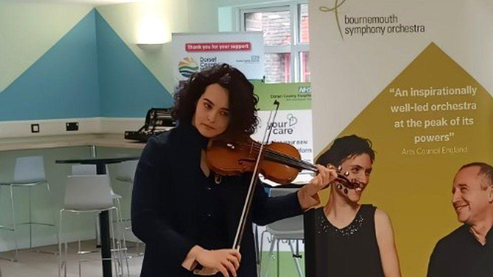 Woman with dark curly hair playing a violin she is wearing a dark coat, large posters and chairs and a desk can be seen behind her