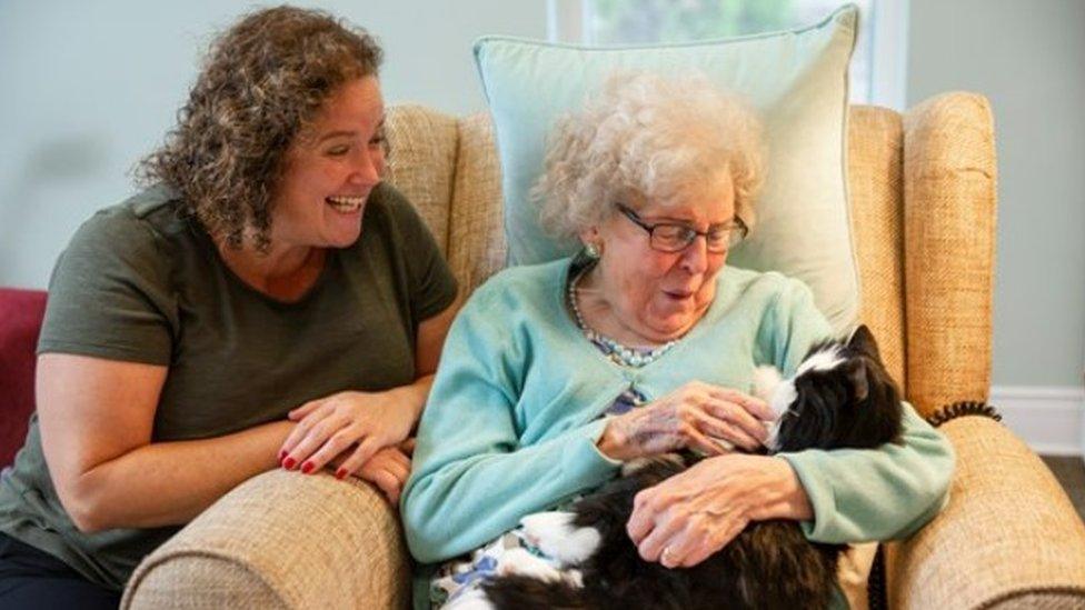 A woman stroking a robotic pet with another woman looking on and smiling