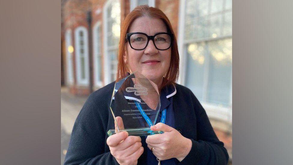 A woman with red hair and black-rimmed glasses smiling at the camera. She is holding a clear trophy and wearing a navy blue nurse's top. There is a brick building behind her.