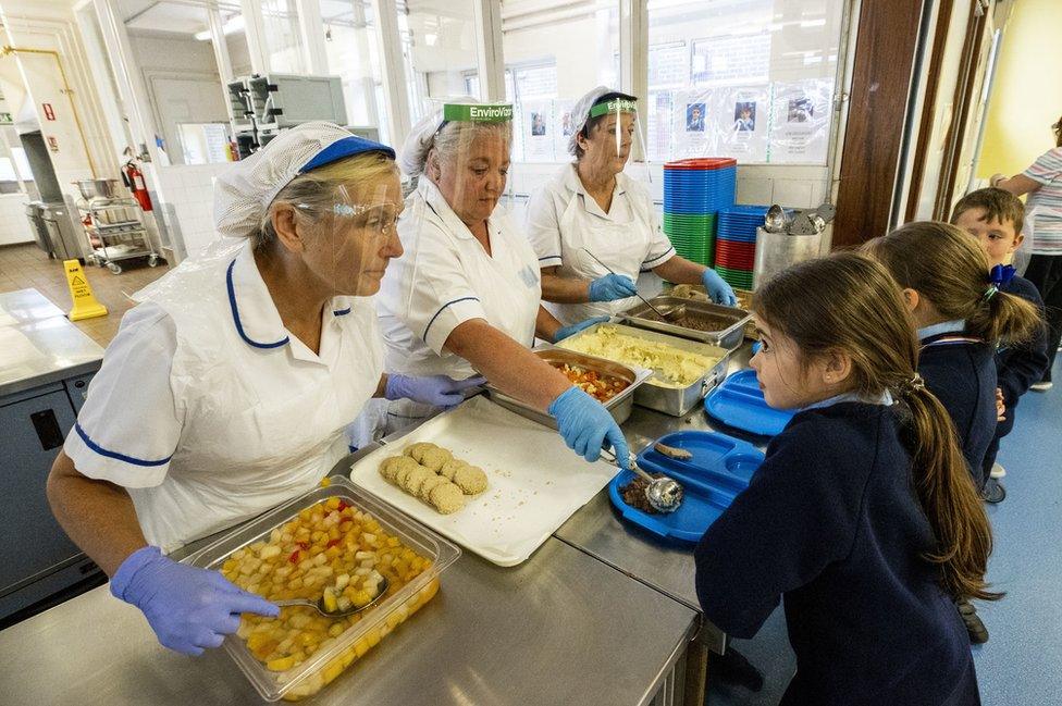 Three dinner-ladies offer lunch to three pupils in a school canteen