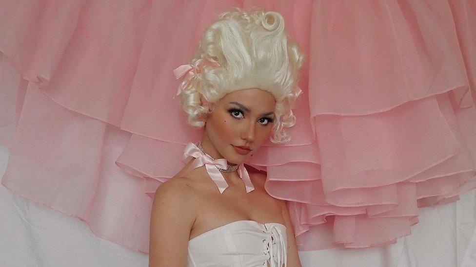 Influencer Janice Glimmer in a white curly wig styled to look like Marie Antionette wearing a corset and a pink bow and jewels around her neck