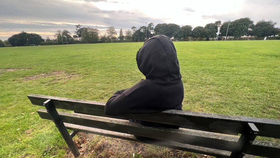 The back of "Amanda" wearing a black hoody sat on a bench looking out over a field