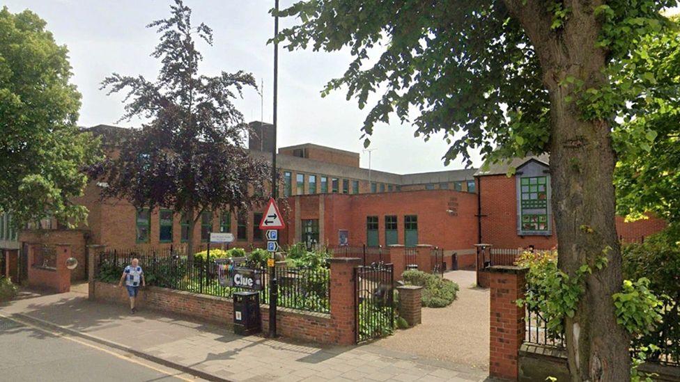 Outside view of a single-storey, red brick former council office in Grantham. In the foreground are green shrubs and green and purple-leaf trees, arranged around a low, red-brick wall with black metal railings..