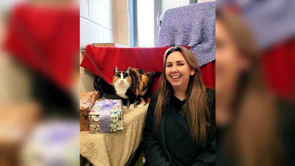 Shelby is smiling and kneeling down next to a cat, which is black, orange and white, and standing on a table next to two wrapped shoeboxes. 