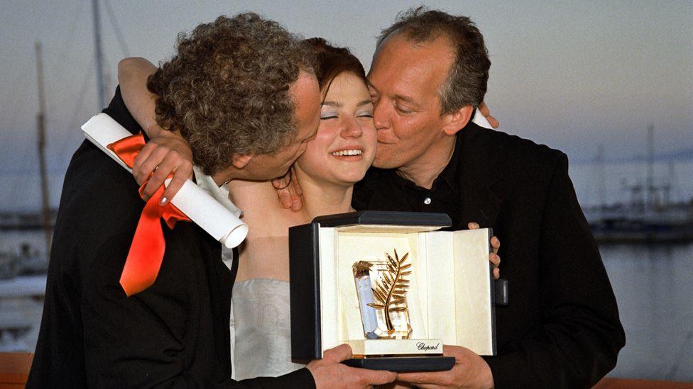 Émilie Dequenne holding a Cannes award with Luc and Jean-Pierre Dardenne on either side kissing her on both cheeks in 1999