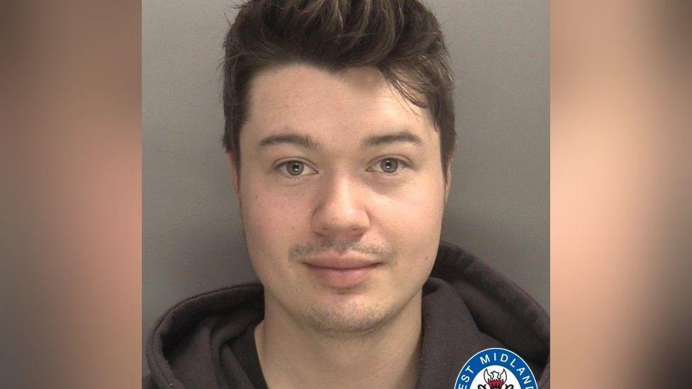 A mugshot of a man with short brown hair, wearing a black hoody