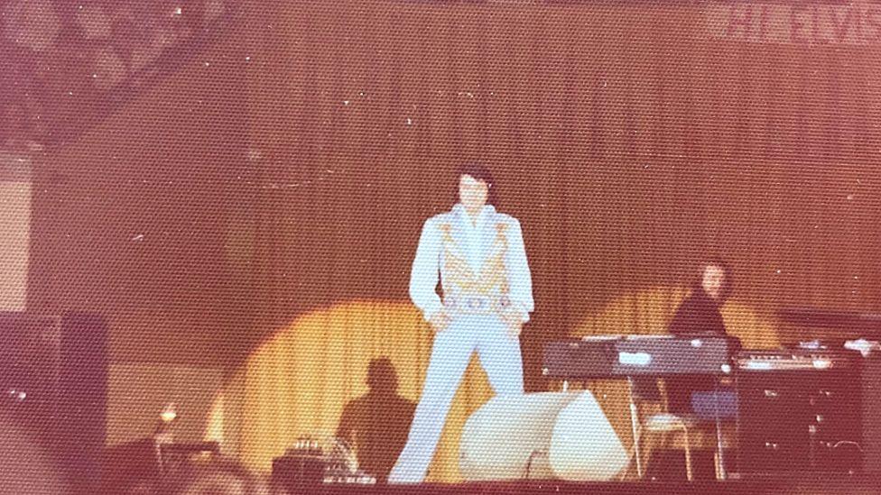 Elvis Presley in concert in 1976. The photo, by Jan Owen, shows Presley dressed in a light-colored jumpsuit with gold embellishments, standing on a stage. The background features a large curtain, and there are musicians and equipment, such as keyboards, on stage to the right. 