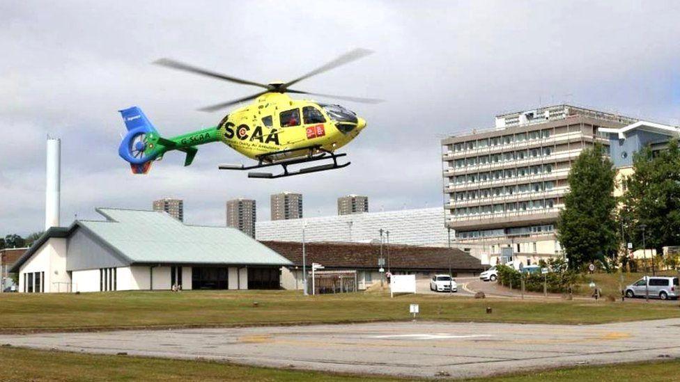 Yellow air ambulance helicopter with green and blue tail, hovering above a heli-pad, with buildings in the background.