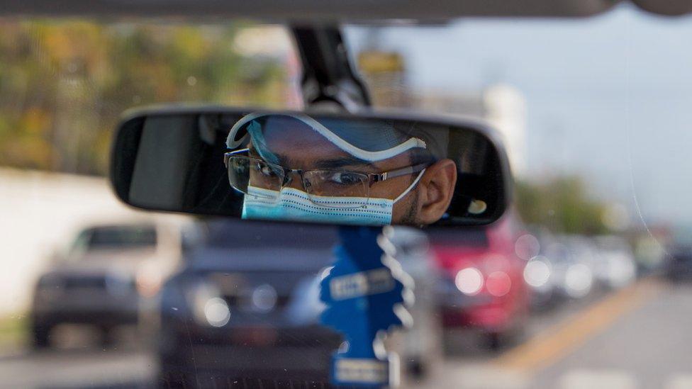 A Uber driver wears a face mask as seen in his rearview mirror