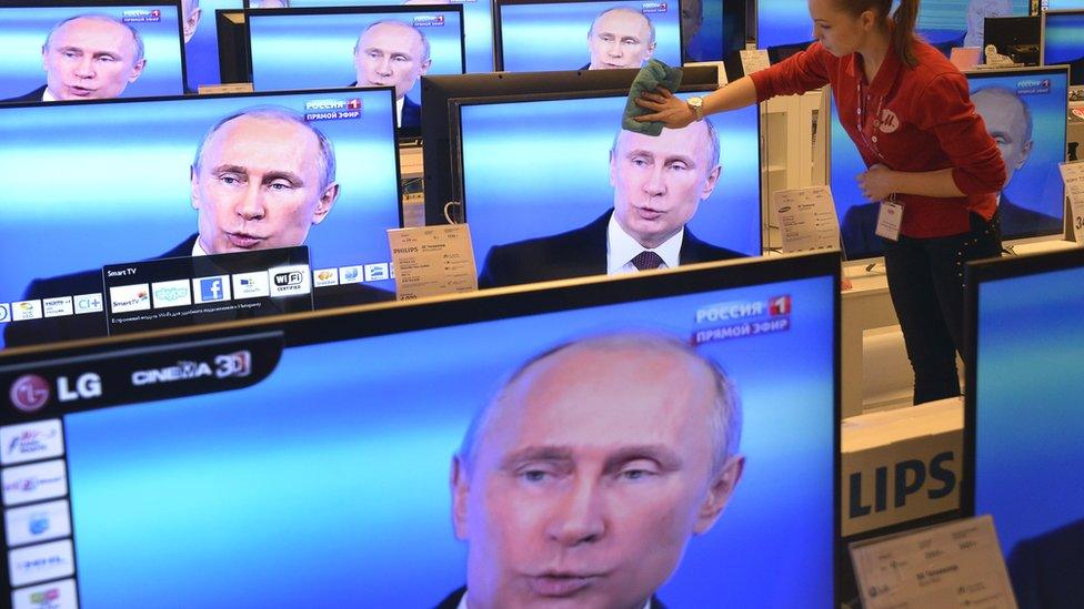 Woman dusts TVs in shop, with Putin on screen