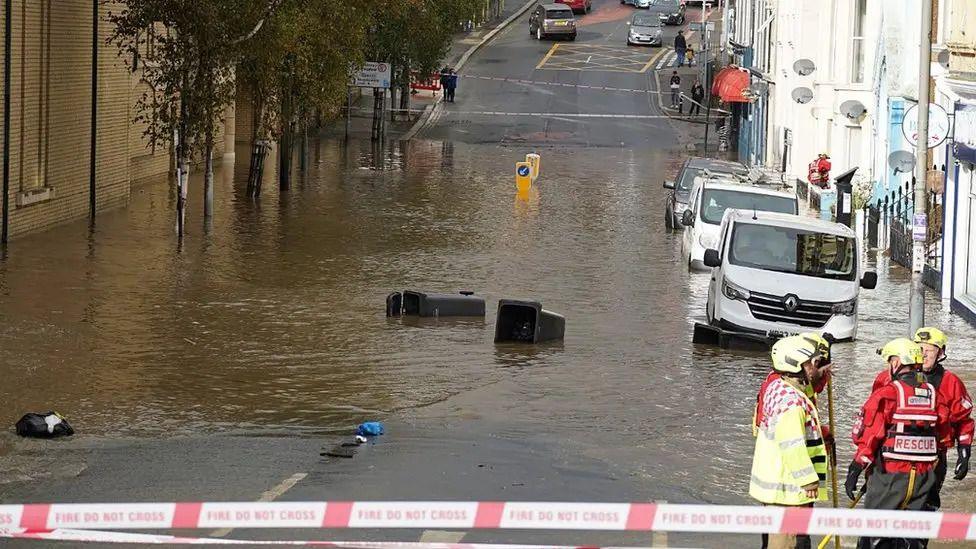 Emergency service crews standing by a large section of road that has been flooded where it dips in the middle. 