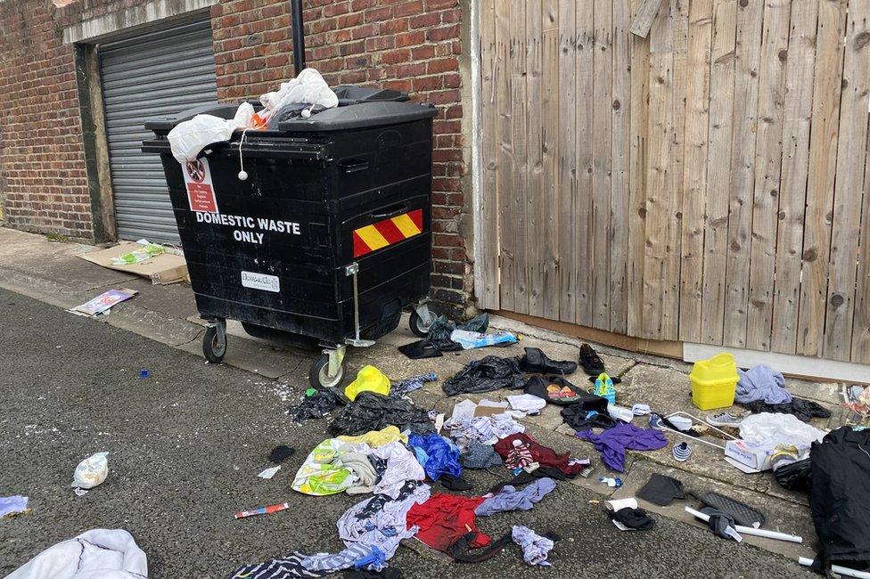 Communal bin with rubbish and clothes on the ground