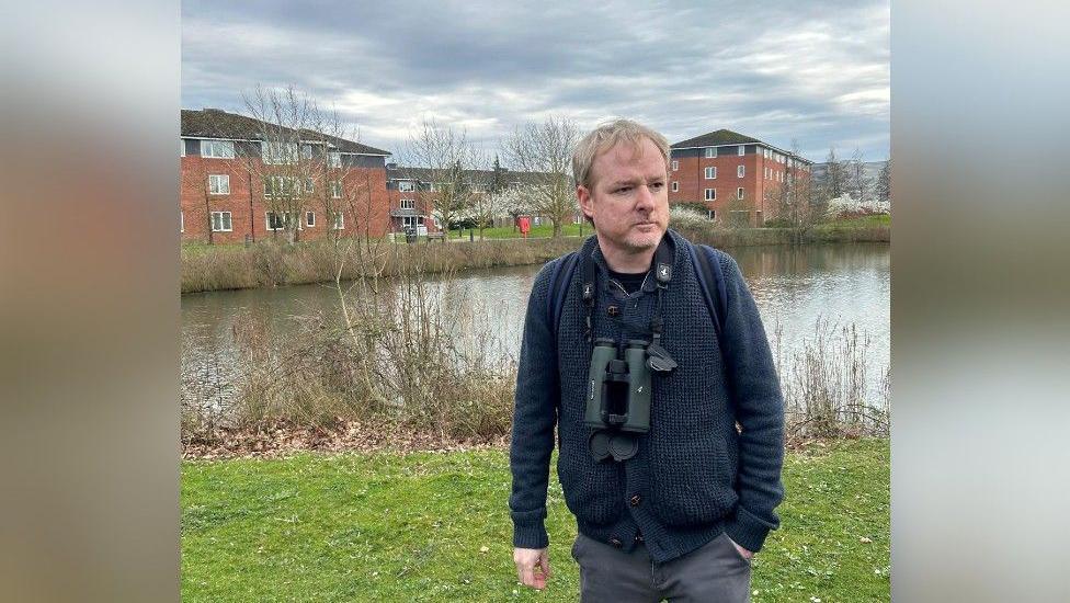 A photo of a man with blond hair, wearing a dark cardigan with a pair of binoculars around his neck. He looks to the right of the camera. He is at the University of Warwick, with grass and a lake behind him. Redbrick buildings are on the other side of the lake.