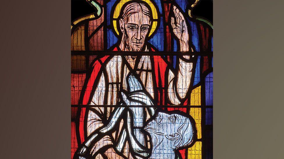 A close-up of a stained glass window showing Christ raising Lazarus from the dead. The bearded Christ, with a golden halo, is wearing a red cloak over a cream robe and looking down towards the white face of Lazarus, who has his head tilted up towards Christ.