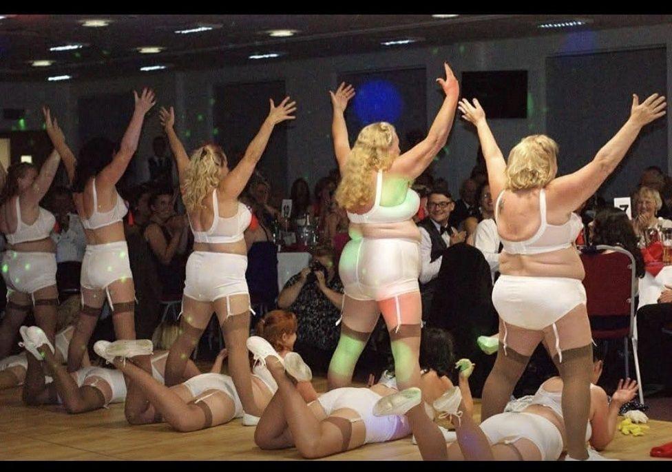 10 women wearing white underwear, stockings and white shoes. Five of the women have their arms outstretched in a V-shape and are standing astride over the other five women who are lying on the floor with their legs bent at the knees and crossed over. They are dancing to a crowd of people who are sat down at round tables.