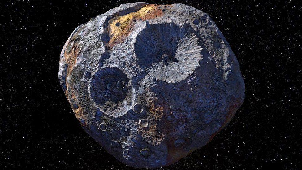 Psyche asteroid, grey and brown with huge craters that appear almost golden in colour nagainst the backdrop of space - black with many white stars.