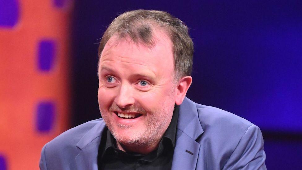 Chris McCausland pictured smiling and wearing a blue jacket and black shirt on a TV studio set.