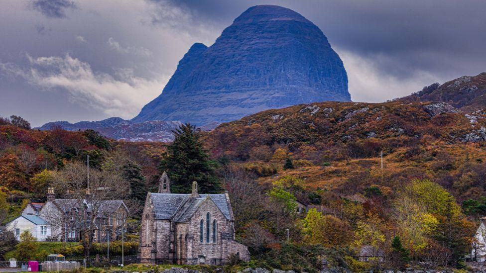 The Suilven mountain towers over a church and house in Lochinver on a cloudy afternoon. The mountain is slightly shrouded by cloud and mist. Orange and brown coloured heather and plants cover the landscape. In the foreground, a church, built with light coloured stone and a dark roof, stands next to a house, which has dark stone and a dark roof. An extension is white with a blue roof.