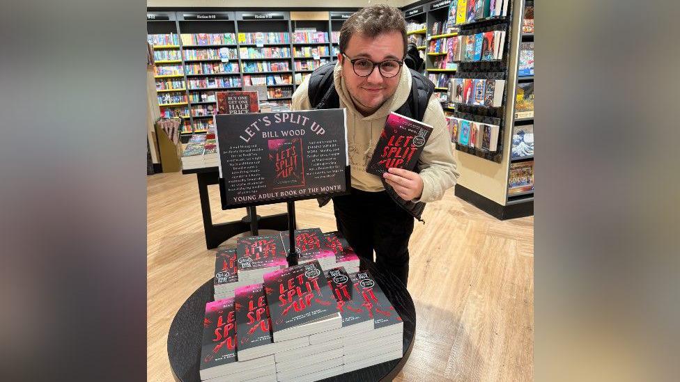 Bill Wood has brown hair and wears black glasses. He is standing next to a stack of his books on a small table. There is a sign that says "Let's split up, Bill Wood". He is holding a copy of his book in his hand. The cover is black with pink/red in the corner