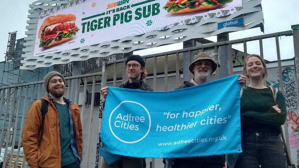 Adblock Bristol protesters next to a billboard. There are 4 of them. 3 protestors are holding a blue Adfree Cities banner, which reads "for happier, healthier cities"