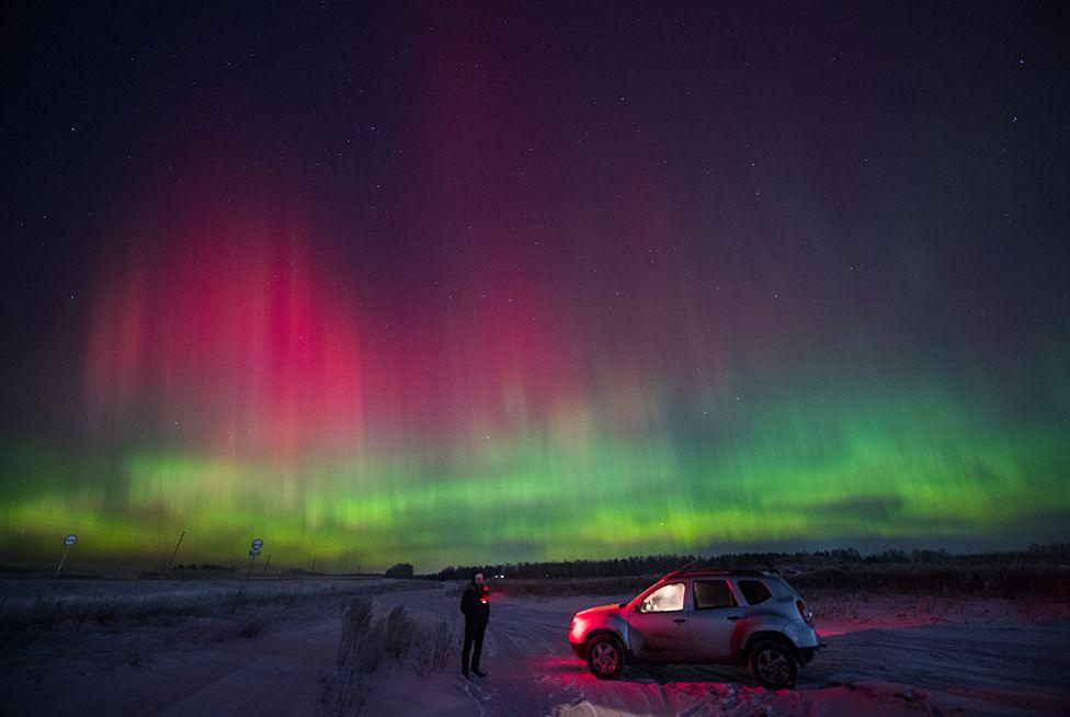 A man, holding a torch, stands next to a vehicle - with the interior light on -to observe the Northern Lights, appearing pink and green in the sky above the Siberian city of Krasnoyarsk, Russia on 2 January, 2025. 